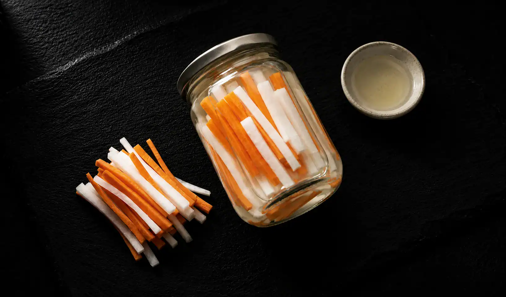 Vietnamese pickled daikon and carrot in a glass jar with rice vinegar on dark slate
