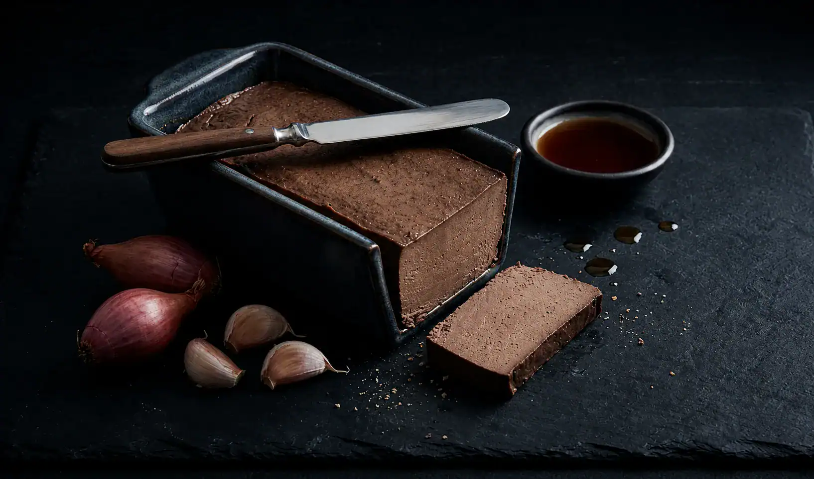 Pork liver pâté in a dark loaf pan with a slice cut beside it, shallots, garlic, and fish sauce on dark slate