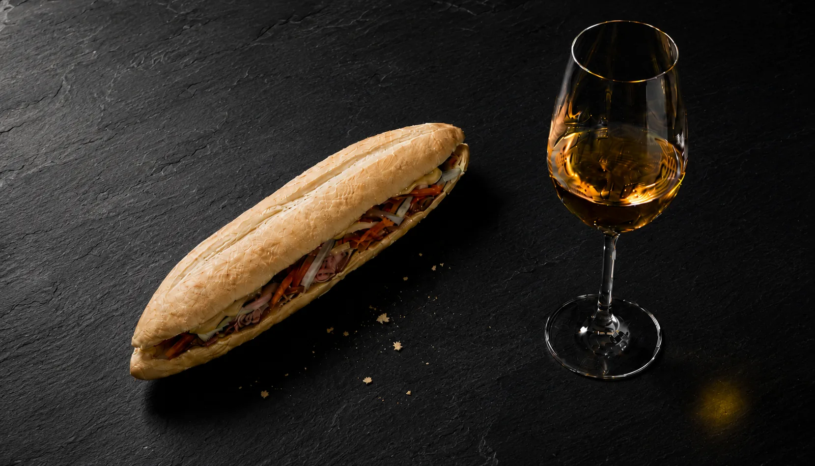 A Vietnamese bánh mì sandwich open on dark slate beside an elegant tulip wine glass holding golden white wine, with natural bread crumbs scattered on the slate